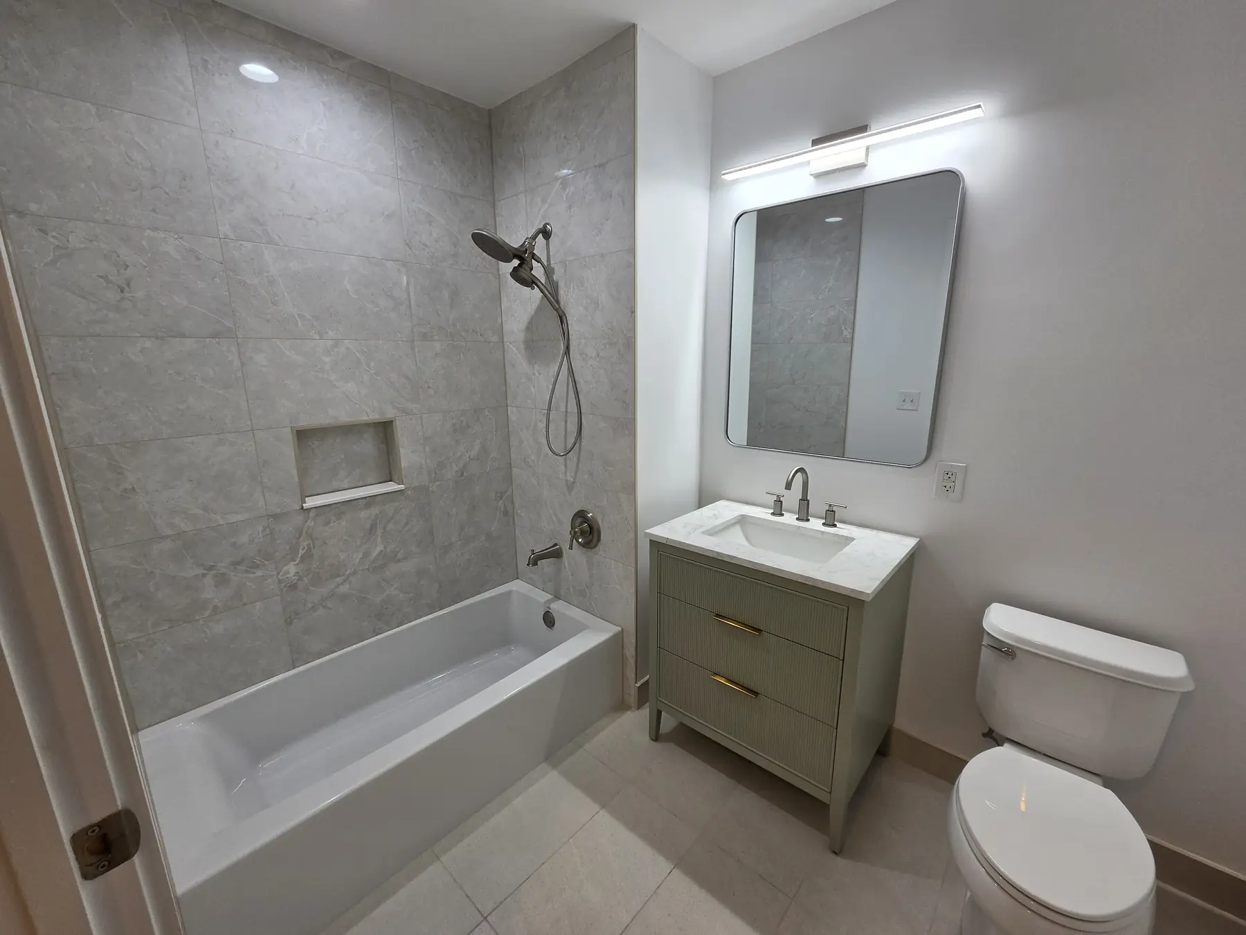 The finished tub bathroom — large-format tile surround with a niche, sage vanity, new mirror and lighting