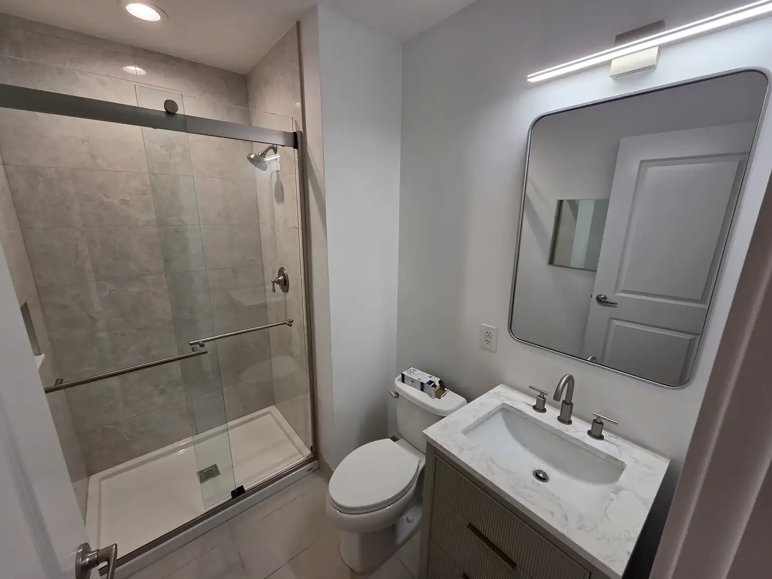 The second bathroom finished — a walk-in shower behind a sliding glass door, with a new vanity and mirror