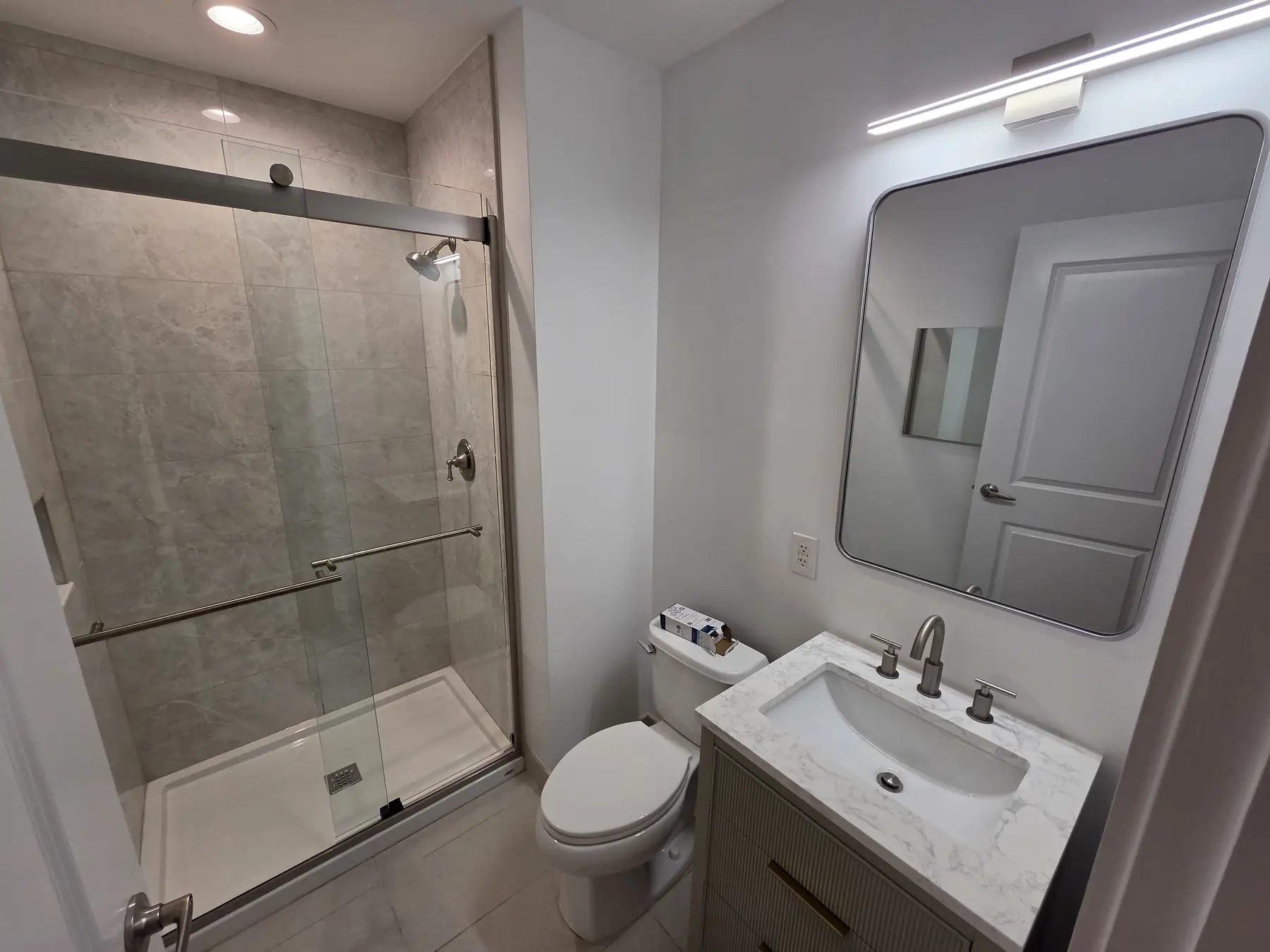 The second bathroom finished — a walk-in shower behind a sliding glass door, with a new vanity and mirror